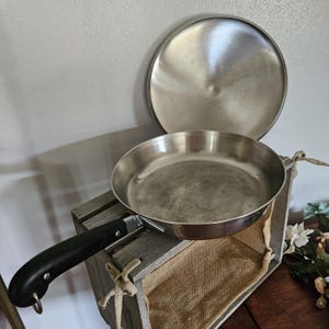May include: Stainless steel frying pan with a black handle and matching lid. The pan is sitting on a wooden crate with a burlap lining. The lid is slightly off to the side, revealing the interior of the pan.