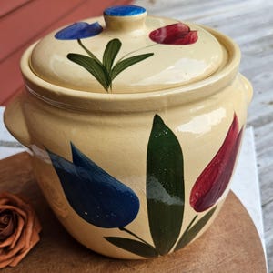 May include: A cream-colored ceramic crock with a matching lid, featuring hand-painted blue and red tulip motifs with green leaves. The round container has two small side handles and a knob on the lid, showcasing a glossy finish throughout.