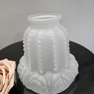 May include: A frosted glass lampshade with a cactus-like design. The shade has a cylindrical shape with vertical beaded detailing and a flared top. The base features a floral pattern. The overall color is white.