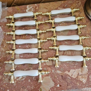 May include: A collection of white and gold cabinet pulls arranged on a marble surface. Each pull features a white, oblong handle with gold-colored decorative accents and mounting hardware. The pulls are likely for furniture or cabinetry.