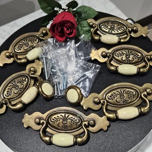 May include: Antique brass cabinet pulls with cream-colored ceramic handles and decorative detailing. The set includes matching knobs and screws for installation. A red rose and greenery are in the background.