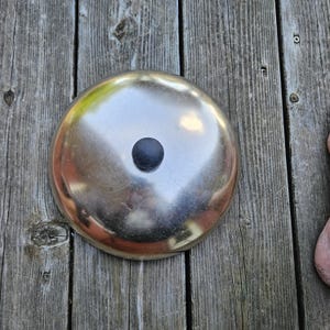 May include: A circular, polished silver-tone metal lid with a smooth, reflective surface. The lid features a small, centered, matte black circular knob handle. The design is simple and functional, with a clean, metallic finish throughout the dome.