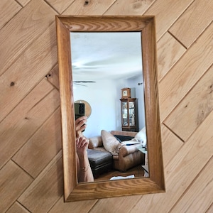 May include: Rectangular wooden framed mirror. The mirror reflects a room with a brown leather couch, a wooden cabinet, and a ceiling fan. The frame is made of light brown wood.