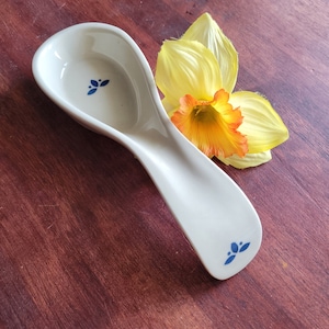 May include: Cream-colored ceramic spoon rest with blue floral accents. The spoon rest has a curved design and is next to a yellow and orange daffodil flower. The spoon rest is on a dark wood surface.