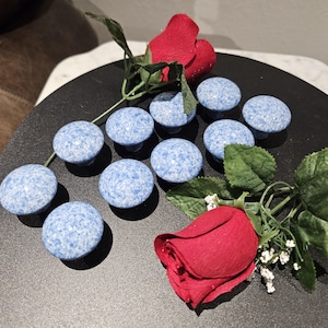 May include: A collection of ten blue and white speckled cabinet knobs arranged on a black surface, with two red roses and green leaves. The knobs have a round shape and a small base for mounting.