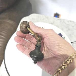 May include: Antique brass and dark-toned cabinet pull with ornate detailing. The pull features a decorative circular backplate, a pivoting arm, and a bulbous handle. This vintage hardware adds a touch of elegance to furniture.