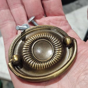 May include: An oval-shaped brass-toned metal drawer pull featuring a central circular medallion with a radiating ribbed pattern, surrounded by a rope-textured border and a hinged drop handle, accompanied by two silver-colored metal mounting screws.