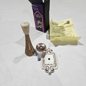 May include: A door stop set from the Sensational Collection, featuring a spring-loaded design with a cream-colored bumper and a brass-colored spring. Includes a decorative white and gold escutcheon plate, a mounting screw, and installation instructions.