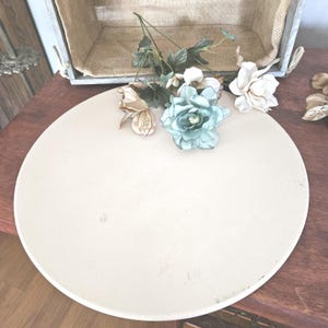 May include: A round, off-white ceramic pizza stone with a few small marks. Artificial flowers in shades of white, gold, and teal are arranged on the stone. A wooden crate with a burlap lining is in the background.