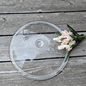 May include: A clear, round glass lid with a central knob and a textured pattern. The lid is resting on a weathered wooden surface, with a sprig of pink and white flowers to the side.
