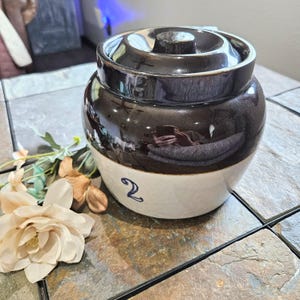May include: A vintage stoneware crock with a dark brown glazed top and a cream-colored base. The crock has a lid and is marked with the number "2" in blue. Decorative flowers are in the foreground.