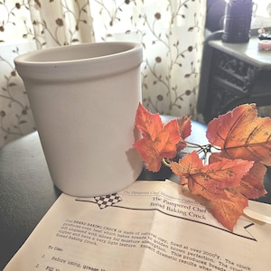 May include: A light-colored stoneware bread baking crock from The Pampered Chef, with a slightly flared rim. The crock is accompanied by a sprig of orange and red autumn leaves and a product description sheet.