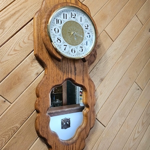 May include: A wall-mounted clock with a wooden frame featuring a scalloped edge. The clock face has black Arabic numerals on a white background with a gold center. Below the clock face is a mirror with a matching scalloped wooden frame.