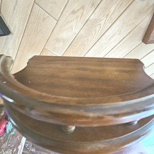 May include: A dark brown, two-tiered wooden side table with a curved front edge and decorative trim. The table has a smooth, polished surface and is likely used for display or storage. The wood grain is visible.
