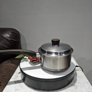 May include: A stainless steel saucepan with a lid and a black handle. The pot is sitting on a white and black rotating display stand. A red rose is placed near the handle.