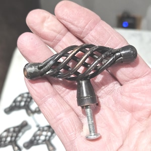 May include: A dark bronze cabinet knob with a cage-like design. The knob features a twisted metal pattern and a threaded screw for installation. Several other similar knobs are visible in the background, suggesting a set for furniture or cabinetry.