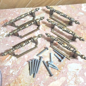 May include: Set of six antique brass drawer pulls with decorative detailing. Each pull has a curved handle and is accompanied by several silver screws for installation. The pulls are arranged on a pink and white marble surface.