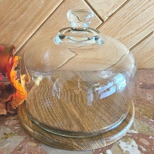 May include: A clear glass cloche with a wooden base and a glass knob on top. The wooden base has a circular shape and a natural wood grain pattern. The cloche is designed to cover and protect items.