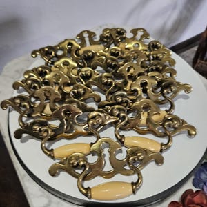 May include: A collection of antique brass drawer pulls with cream-colored handles. The pulls have an ornate, scrolled design and are arranged on a white surface. The brass has a warm, aged patina, suggesting vintage hardware.