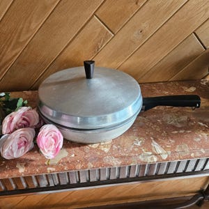May include: A vintage silver-colored pancake griddle with a black handle and a black knob on the lid. The griddle sits on a red and white marble surface, with pink roses to the left. The griddle is a kitchenware item.
