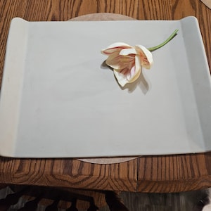 May include: A rectangular, light gray ceramic serving tray with slightly raised, curved edges on two sides. A single artificial flower with white petals featuring delicate pink and red streaks and a green stem rests on the tray surface.