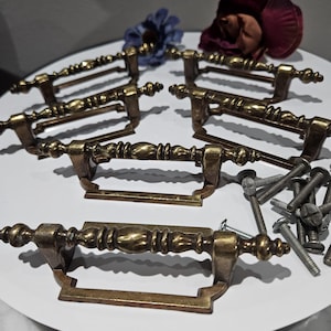 May include: Six antique brass drawer pulls with ornate detailing. Each pull features a decorative handle and a rectangular base. Several silver screws are included. The pulls are arranged on a white surface with a few decorative flowers.