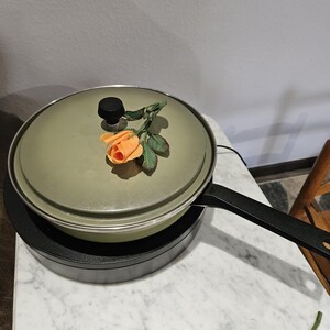 May include: A green frying pan with a lid and a black handle. An orange rose and green leaves rest on the lid. The pan sits on a black heating element, and the handle extends to the right.