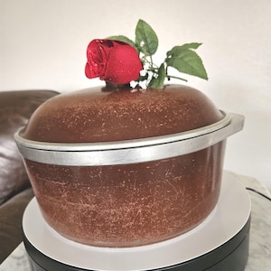 May include: A brown vintage cooking pot with a lid and silver trim. A red rose with green leaves and white accents sits atop the lid. The pot is on a white and black surface.