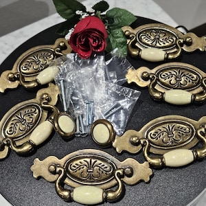 May include: A set of five antique brass-toned metal drawer pulls featuring oval backplates with embossed floral patterns and cream-colored ceramic handles. The set includes a small round knob and several metal screws in clear plastic bags.