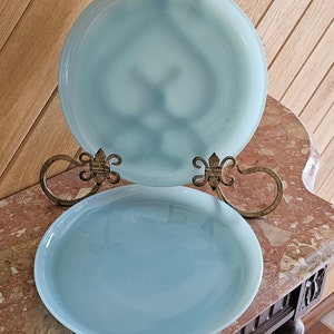 May include: Two light blue glass plates displayed on decorative gold-colored metal stands. The plates are round and have a glossy finish. The stands have a fleur-de-lis design.