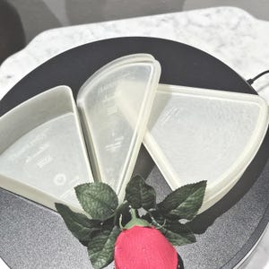 May include: Three clear, triangular food storage containers with lids are arranged on a black surface. A red rose with green leaves is in the foreground. The containers appear to be made of plastic.