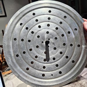 May include: A circular, silver-colored metal steaming rack featuring a series of concentric rings and evenly spaced perforations throughout the surface. A small, dark, cylindrical handle is positioned at the center of the disc to facilitate lifting.