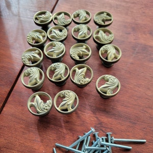 May include: A collection of antique-style brass cabinet knobs with a floral design. The knobs are round with a raised floral relief within a circular frame. Several screws are visible at the bottom of the image. The knobs are arranged on a dark wood surface.
