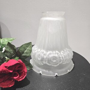 May include: A frosted glass lampshade with a flared shape. The lampshade features vertical lines and a band of embossed floral designs around the base. The shade is white and sits on a dark surface.