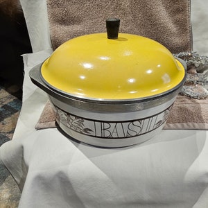 May include: A vintage yellow lidded casserole dish with a black handle. The white body features a brown floral design and the word "BASIL" in large letters. The dish has a silver rim.
