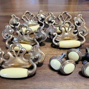 May include: A collection of three ornate brass drawer pulls with cream-colored ceramic handles and four matching round brass knobs with cream-colored ceramic centers. The brass hardware features a decorative, scrolled backplate design.