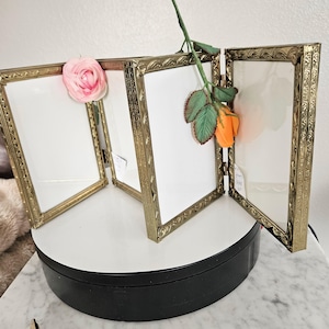 May include: A gold-toned, four-panel picture frame with ornate detailing. Each panel holds a white insert. A pink and orange rose are placed in the frame. The frame is on a white surface.