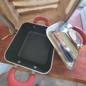May include: A black square cooking pan with red handles and a silver lid. The pan has a non-stick surface and is sitting on a wooden table. The lid is partially open, reflecting the surroundings.
