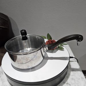 May include: A stainless steel saucepan with a glass lid and a black handle. The pan is sitting on a white surface, with a red rose and green leaves next to it. The handle has a metal ring at the end.