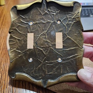 May include: A rectangular brass light switch plate with a double toggle configuration. The surface features a dark, textured, crinkled finish with raised edges and four circular screw holes positioned at the corners of the switch openings.