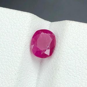 Könnte beinhalten: Ein facettierter, oval geformter Rubin-Edelstein. Der Edelstein hat eine tiefe, satte rote Farbe mit einer polierten Oberfläche, die das Licht reflektiert. Der Rubin liegt vor einem weißen Hintergrund.