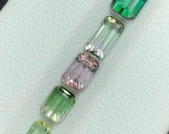 Puede incluir: Una variedad de gemas de corte rectangular en tonos verdes y rosas. Las gemas están dispuestas en diagonal, mostrando sus facetas pulidas y claridad interna. Las gemas se exhiben sobre una superficie blanca.