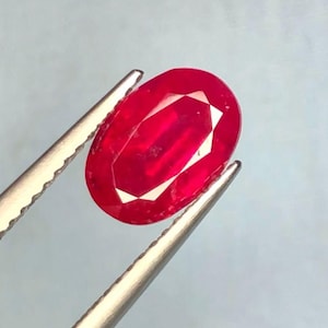 Puede incluir: Una gema ovalada de color rojo vibrante sostenida por unas pinzas. La gema está facetada y tiene un alto pulido.
