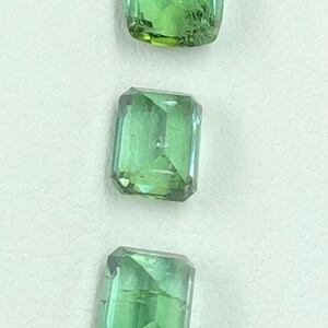 May include: Three green gemstones with a rectangular shape and faceted edges are shown against a white backdrop. The gems are translucent, allowing light to pass through, and display a range of green hues.