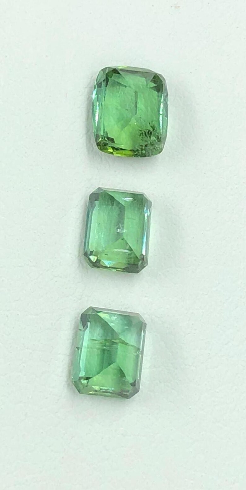 May include: Three green gemstones with a rectangular shape and faceted edges are shown against a white backdrop. The gems are translucent, allowing light to pass through, and display a range of green hues.