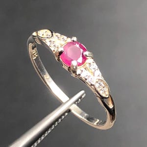 May include: A silver ring featuring a central round-cut red ruby gemstone held by four prongs. The band is accented with small clear stones set along the shoulders, and the interior of the band is stamped with the hallmark 925 for sterling silver.