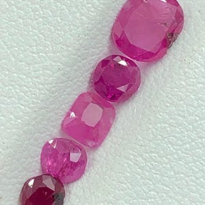 Peut inclure: Une collection de six pierres précieuses rose-rouge taillées, de formes et de tailles variées. Les pierres précieuses sont disposées en diagonale, mettant en valeur leurs surfaces polies et leur clarté interne. Les gemmes sont posées sur un fond blanc texturé.