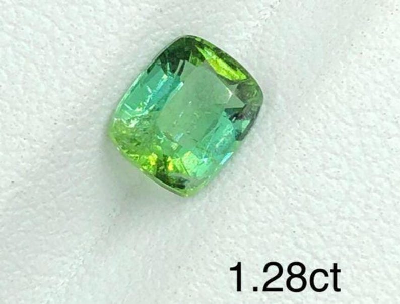 May include: A faceted, cushion-cut gemstone displaying a mix of green and teal colours. The gem is set against a white backdrop. The text "1.28ct" is visible, indicating the carat weight of the stone.