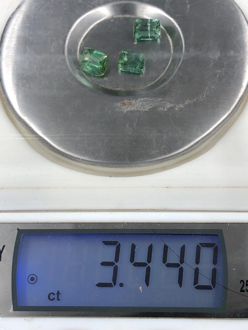 May include: Three faceted, emerald-green gemstones are displayed on a silver scale. The digital display shows a weight of 3.440 ct. The gemstones are rectangular and appear to be of similar size and shape.