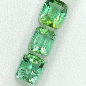 May include: Three faceted, green gemstones with a rectangular shape. The gems have a vibrant, light green colour with a slight yellow hue. The gemstones are displayed on a white surface.